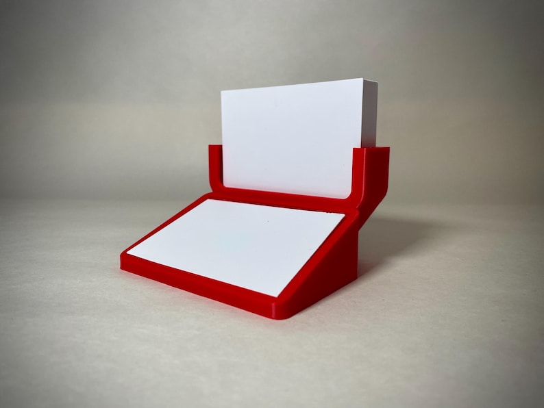 May include: A red and white business card holder. The holder has a red frame with a white surface for the cards and a white block to hold the cards in place. The design is modern and minimalist.