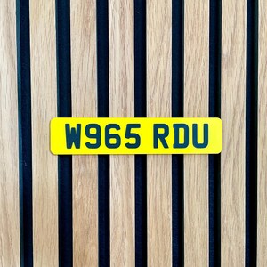 May include: A bright yellow license plate with the text "W965 RDU" in dark blue lettering. The plate is set against a backdrop of vertical wooden slats with dark blue stripes.