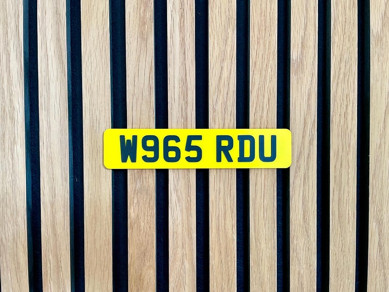 May include: A bright yellow license plate with the text "W965 RDU" in dark blue lettering. The plate is set against a backdrop of vertical wooden slats with dark blue stripes.