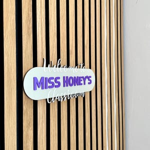 K&ouml;nnte beinhalten: Ein Klassenzimmerschild mit dem Text "Welcome to Miss Honey's Classroom" in Wei&szlig; und Lila. Das Schild ist an einer Holzwand mit vertikalen schwarzen und braunen Streifen angebracht.