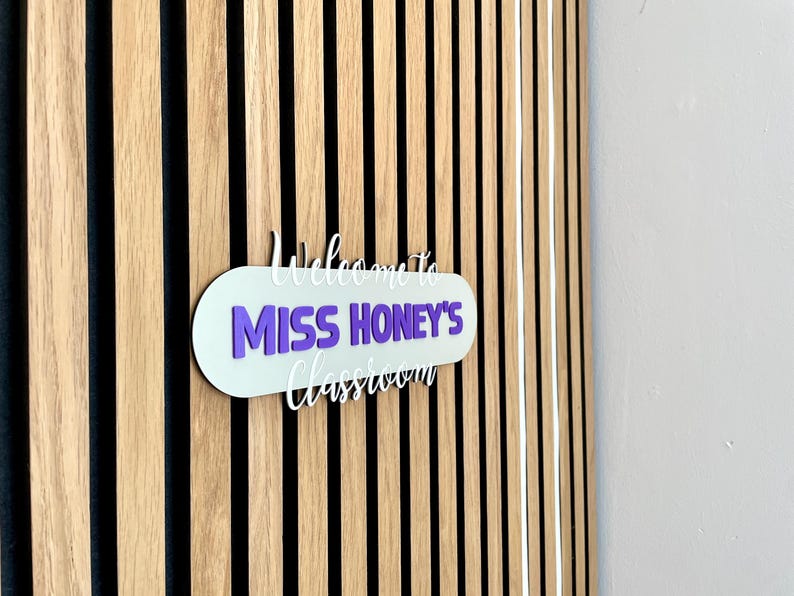 K&ouml;nnte beinhalten: Ein Klassenzimmerschild mit dem Text "Welcome to Miss Honey's Classroom" in Wei&szlig; und Lila. Das Schild ist an einer Holzwand mit vertikalen schwarzen und braunen Streifen angebracht.