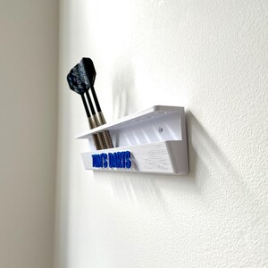 May include: A white wall-mounted dart holder with three darts. The holder has a shelf and a lower section with the blue text "TOM'S DARTS". The darts have black shafts and silver tips. The holder is mounted on a white textured wall.