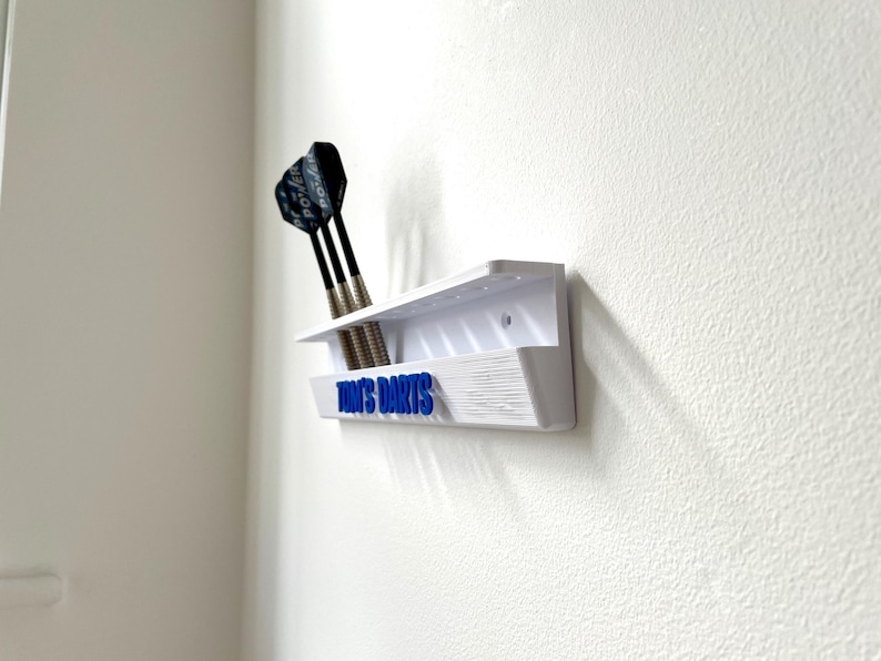 May include: A white wall-mounted dart holder with three darts. The holder has a shelf and a lower section with the blue text "TOM'S DARTS". The darts have black shafts and silver tips. The holder is mounted on a white textured wall.