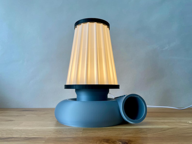 Puede incluir: Una l&aacute;mpara de mesa azul y blanca con una pantalla de tela plisada. La base de la l&aacute;mpara tiene forma de turbina.