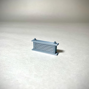 May include: A small, gray, rectangular decorative fence with horizontal slats. The fence has decorative posts on each end and is likely a miniature model or architectural element. The object is set against a plain, light background.