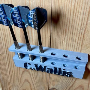 Silver / grey wall mounted dart holder with large black text along the lower edge showing a name. 3 darts in the holder with a total of 6 available slots. Shown mounted to a wooden background.