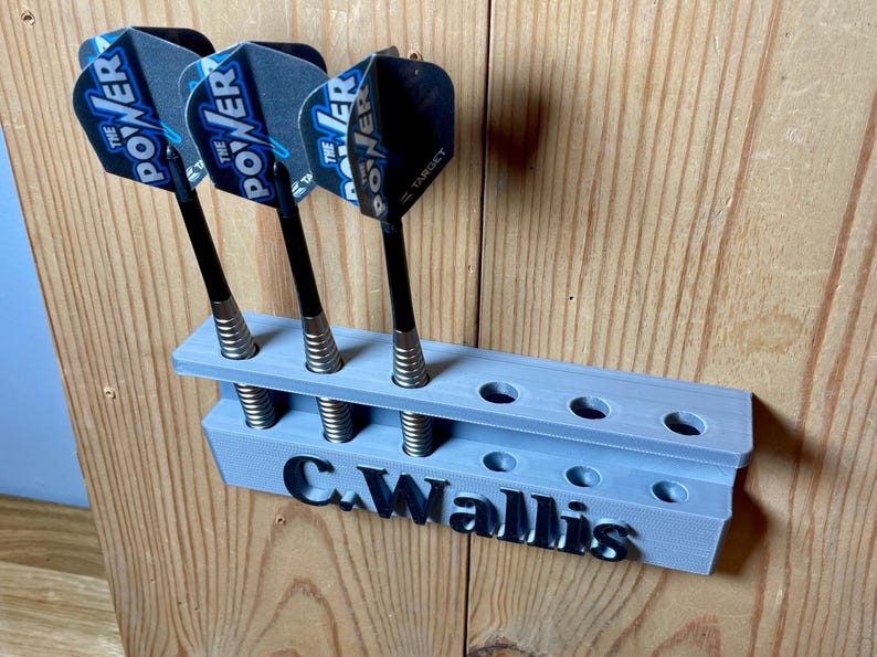 Silver / grey wall mounted dart holder with large black text along the lower edge showing a name. 3 darts in the holder with a total of 6 available slots. Shown mounted to a wooden background.