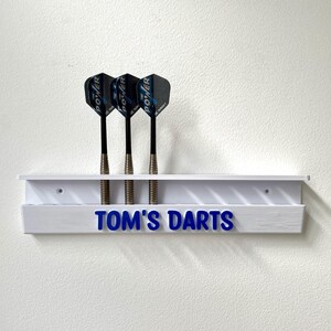 May include: A white dart holder with three darts. The darts have black shafts and blue and black flights. The holder has the text "TOM'S DARTS" in blue.