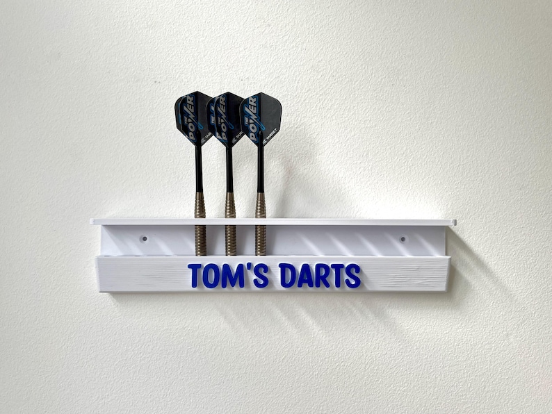 May include: A white dart holder with three darts. The darts have black shafts and blue and black flights. The holder has the text "TOM'S DARTS" in blue.