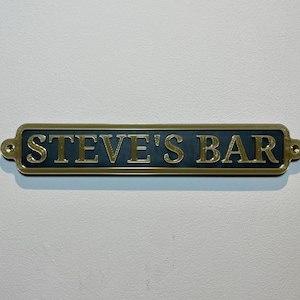 May include: A gold-colored metal sign with black lettering that reads "STEVE'S BAR".