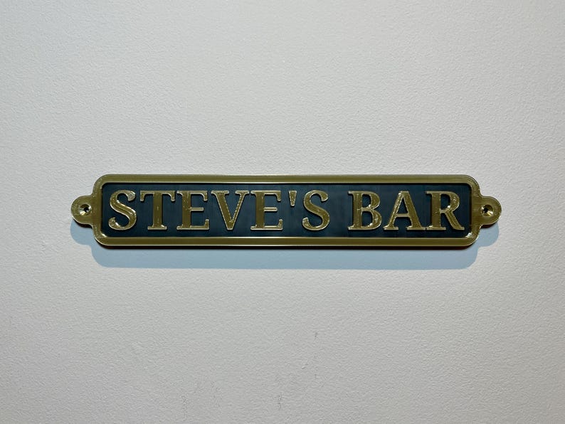 May include: A gold-colored metal sign with black lettering that reads "STEVE'S BAR".