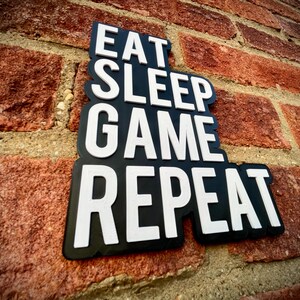 May include: A black sign with the words "EAT SLEEP GAME REPEAT" in white, mounted on a red brick wall. The sign has a unique, layered design, and the text is in a bold, easy-to-read font. The brick wall provides a textured background.