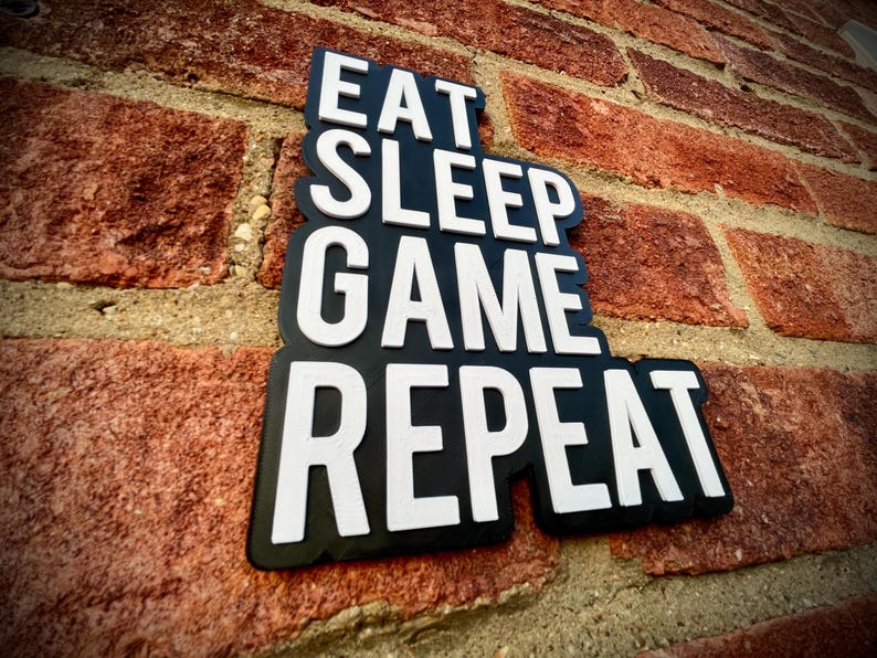 May include: A black sign with the words "EAT SLEEP GAME REPEAT" in white, mounted on a red brick wall. The sign has a unique, layered design, and the text is in a bold, easy-to-read font. The brick wall provides a textured background.