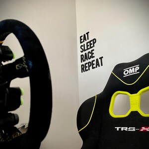 May include: Black racing simulator seat with yellow accents and the text "TRS-X". A black steering wheel with yellow details is visible on the left. The wall has the text "EAT SLEEP RACE REPEAT".