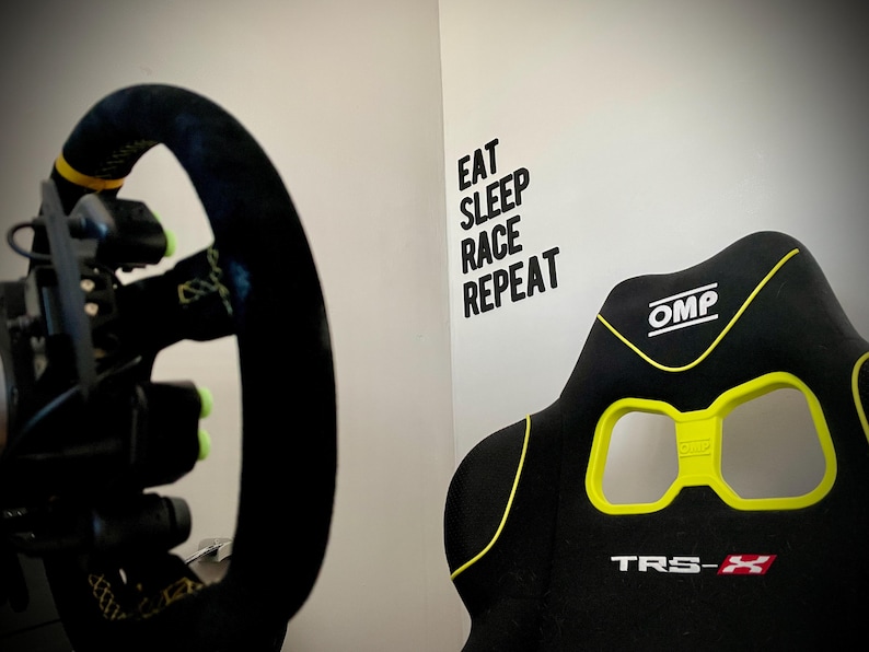 May include: Black racing simulator seat with yellow accents and the text "TRS-X". A black steering wheel with yellow details is visible on the left. The wall has the text "EAT SLEEP RACE REPEAT".