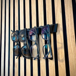 May include: A black wall-mounted glasses rack holding five pairs of eyeglasses and sunglasses. The glasses are black-framed and hang against a backdrop of vertical wooden slats in alternating light and dark tones.