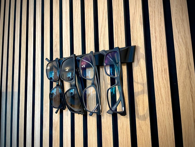 May include: A black wall-mounted glasses rack holding five pairs of eyeglasses and sunglasses. The glasses are black-framed and hang against a backdrop of vertical wooden slats in alternating light and dark tones.