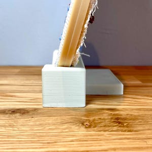 May include: A white, 3D printed stand with a rectangular base and a smaller rectangular platform on top. The stand is designed to hold a circular embroidery hoop.