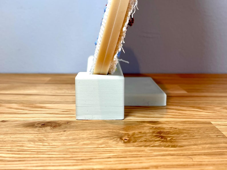 May include: A white, 3D printed stand with a rectangular base and a smaller rectangular platform on top. The stand is designed to hold a circular embroidery hoop.