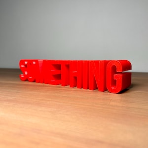 Pu&ograve; includere: Un'insegna tridimensionale rosso brillante con la scritta "SOMETHING". Le lettere sono a blocchi e si ergono su una superficie di legno. L'insegna &egrave; un pezzo d'accento vibrante.
