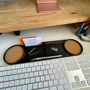 Może przedstawiać: Czarny organizer na biurko z dwiema okrągłymi wkładkami korkowymi, centralną tacką do przechowywania oraz nadrukowanym na pomarańczowo napisem "Layered Shape HQ". Organizer zawiera wizyt&oacute;wkę Toma Ebbsa, dwa małe metalowe narzędzia, pamięć USB i n&oacute;ż introligatorski.