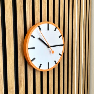 May include: A round orange clock with black hands and black markings on a white face. The clock is mounted on a wall with vertical wooden slats.