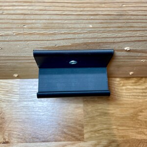 May include: A black metal bracket with a single screw in the center. The bracket is mounted to a wooden surface.