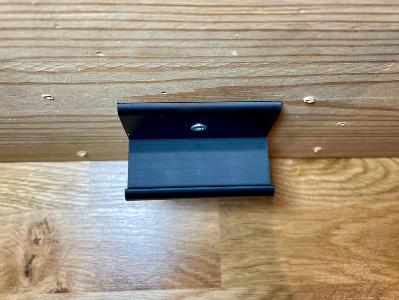 May include: A black metal bracket with a single screw in the center. The bracket is mounted to a wooden surface.