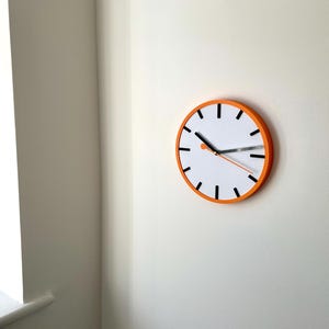 May include: A white clock face with black hour and minute markings and black hands. The clock has an orange rim and is mounted on a white wall.