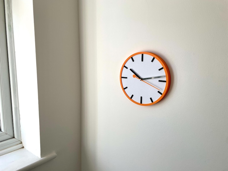 May include: A white clock face with black hour and minute markings and black hands. The clock has an orange rim and is mounted on a white wall.