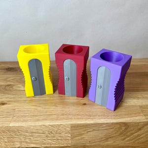 K&ouml;nnte beinhalten: Drei Bleistiftspitzer in verschiedenen Farben: Gelb, Rot und Lila. Jeder Spitzer hat ein graues Messer und ein geripptes Design.