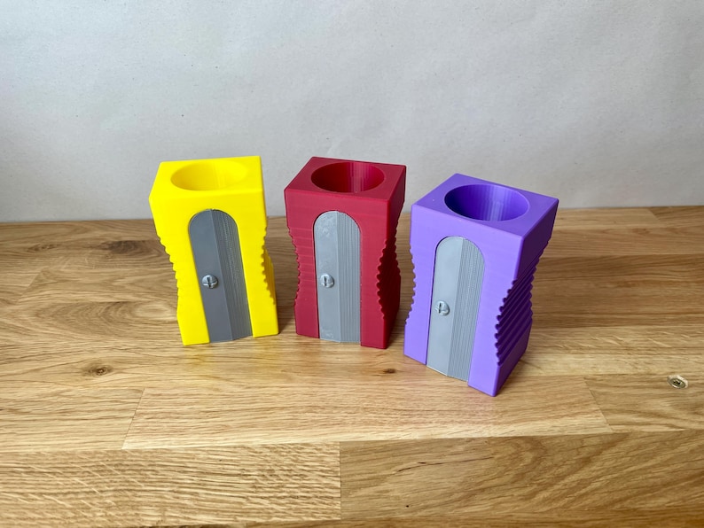 K&ouml;nnte beinhalten: Drei Bleistiftspitzer in verschiedenen Farben: Gelb, Rot und Lila. Jeder Spitzer hat ein graues Messer und ein geripptes Design.