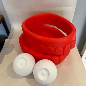 May include: A bright red, large, open-mouthed sculpture with two white spherical eye components. The sculpture is made of a solid material and sits on a neutral surface. The design is reminiscent of a cartoon character.