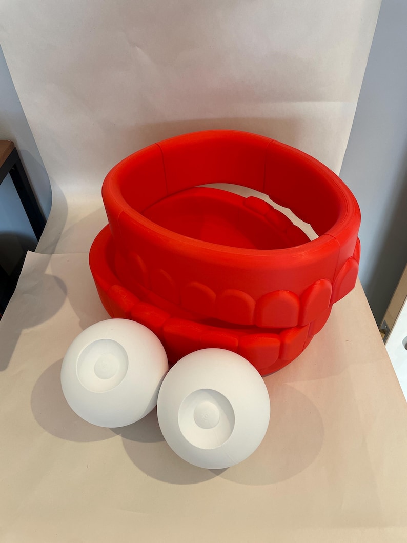 May include: A bright red, large, open-mouthed sculpture with two white spherical eye components. The sculpture is made of a solid material and sits on a neutral surface. The design is reminiscent of a cartoon character.