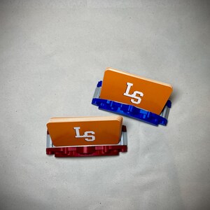 May include: Two orange card holders with the letters "LS" in white. One holder has a blue base, and the other has a red base. The holders are on a light gray surface.