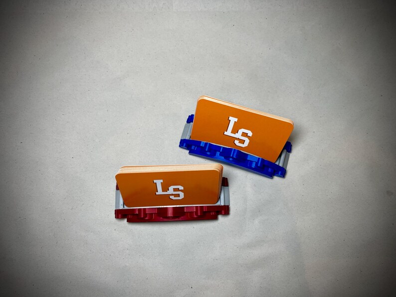 May include: Two orange card holders with the letters "LS" in white. One holder has a blue base, and the other has a red base. The holders are on a light gray surface.