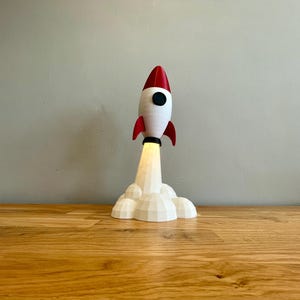Op de afbeelding: Een witte en rode raketlamp met een zwart patrijspoort en rode vinnen. De raket lanceert vanaf een witte wolkenbasis, met een warm licht dat van onderaf uitstraalt. De lamp staat op een houten oppervlak.