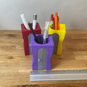 K&ouml;nnte beinhalten: Drei Stiftehalter, einer rot, einer gelb und einer lila, mit einem integrierten Bleistiftspitzer im lilafarbenen Halter. Die Halter stehen auf einer Holzoberfl&auml;che.
