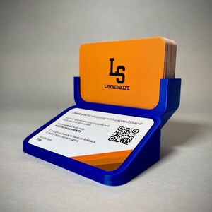 May include: A blue business card holder with a stack of orange cards. The top card displays the black logo "LS" above "LAYEREDSHAPE". The base has a white card with a QR code and text.