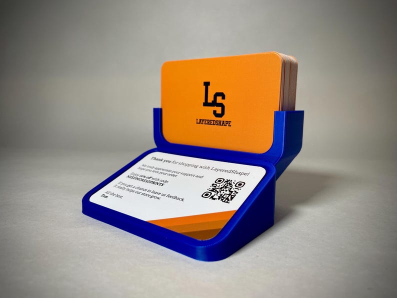 May include: A blue business card holder with a stack of orange cards. The top card displays the black logo "LS" above "LAYEREDSHAPE". The base has a white card with a QR code and text.