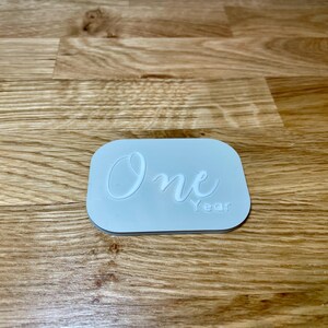 May include: A light blue rectangular sign with the word "One" written in white cursive script and the word "Year" written in white below it.