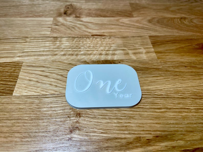 May include: A light blue rectangular sign with the word "One" written in white cursive script and the word "Year" written in white below it.