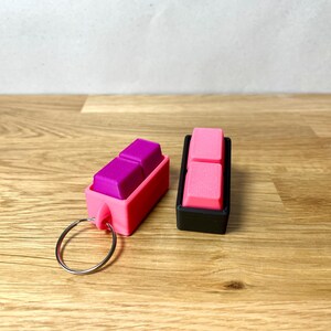 May include: Two keychains with pink and black cases. The keychain on the left has a purple keycap inside. The keychain on the right has a pink keycap inside.