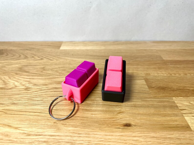 May include: Two keychains with pink and black cases. The keychain on the left has a purple keycap inside. The keychain on the right has a pink keycap inside.
