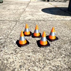 May include: Five orange and grey traffic cones with black bases are arranged on a concrete surface. The cones have a reflective silver band. The image is taken in natural light, with the sun casting shadows.