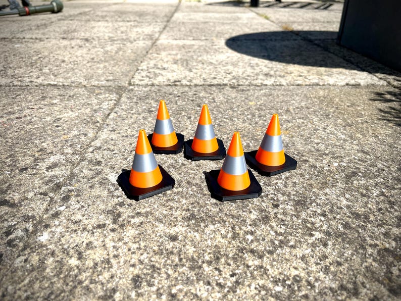 May include: Five orange and grey traffic cones with black bases are arranged on a concrete surface. The cones have a reflective silver band. The image is taken in natural light, with the sun casting shadows.