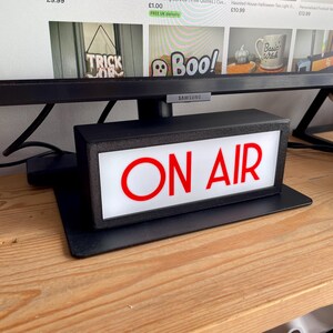 May include: A black rectangular "ON AIR" sign with red lettering and a white background. The sign is on a black base and sits on a wooden surface. The sign is illuminated and appears to be plugged in.
