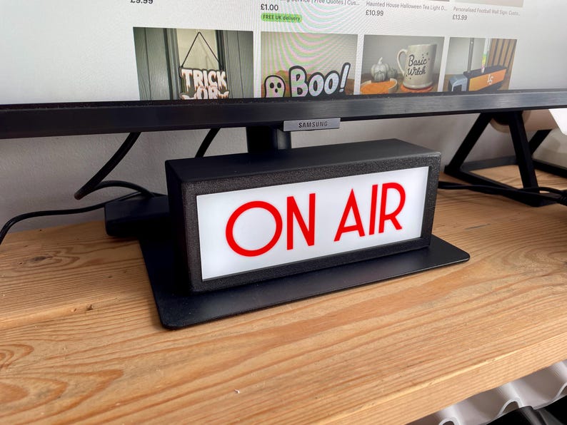 May include: A black rectangular "ON AIR" sign with red lettering and a white background. The sign is on a black base and sits on a wooden surface. The sign is illuminated and appears to be plugged in.