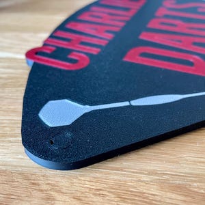Triangular darts sign with the text charlie's darts lounge in red. Main background is black and has silver inlayed darts logos.  Close up