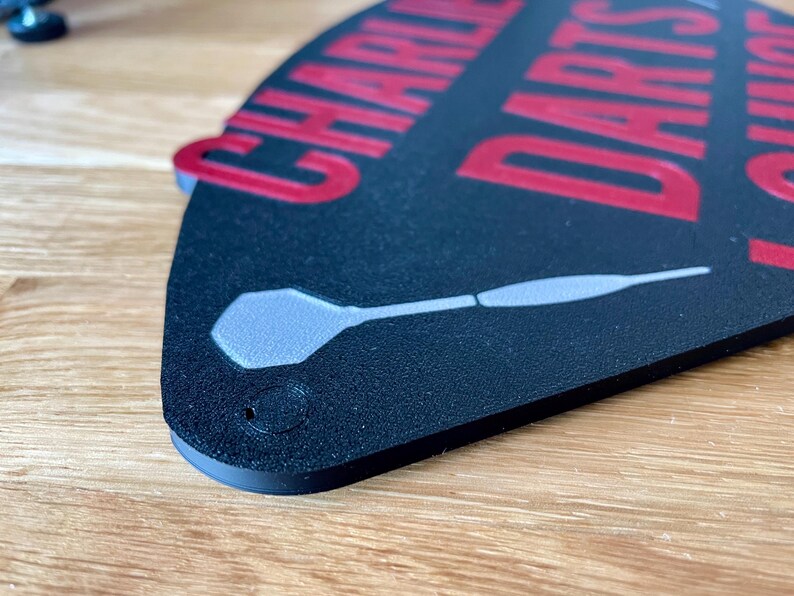 Triangular darts sign with the text charlie's darts lounge in red. Main background is black and has silver inlayed darts logos.  Close up